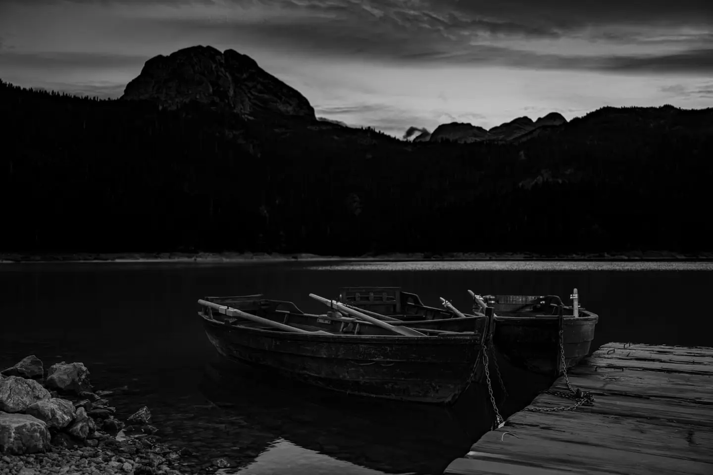 Two weathered wooden rowboats rest chained to a simple plank dock on the still surface of Black Lake (Crno jezero) in Montenegro's Durmitor National Park.
