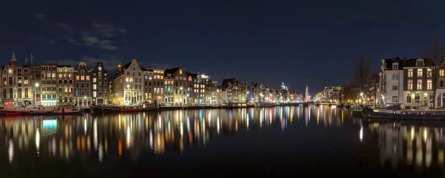 A glass-still Amsterdam canal at night mirrors a long row of historic gabled houses on both banks, with the lit Munttoren rising in the distant centre of the frame.