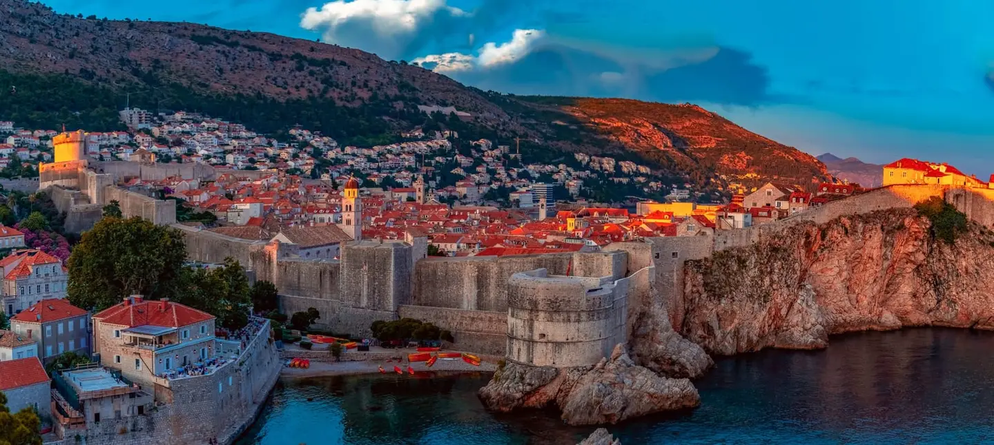 The last light of day washes the medieval ramparts and terracotta rooftops of Dubrovnik old city in warm amber from across the cliff-side bay.