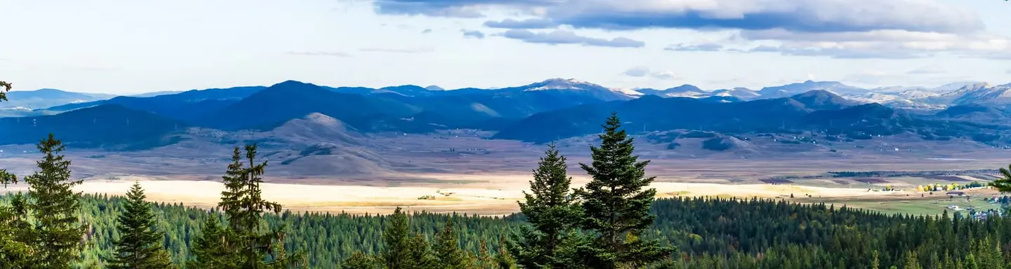 An end-of-day view from the wooded edge of Durmitor National Park out across a wide pastoral basin to a long blue ridge of distant peaks.
