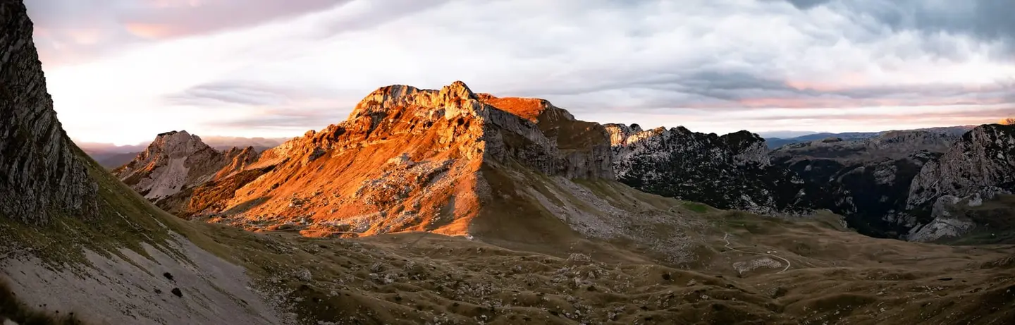 First-light gold rakes across the broad limestone ridge of the Durmitor massif in northern Montenegro, picking out the orange flank of the central peak against deeper-shadowed flanks on either side.