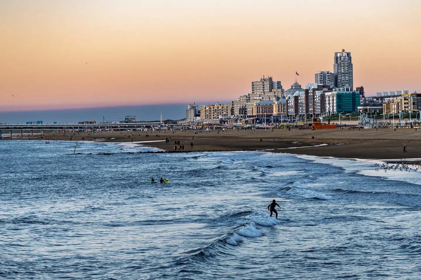 A lone surfer rides a small wave as the pastel sunset glows behind the seaside skyline of Scheveningen.