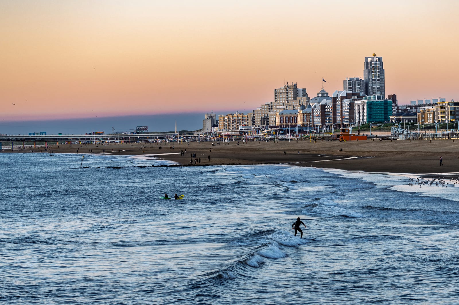 Een eenzame surfer glijdt over een kleine golf terwijl het pastelkleurige avondrood oplicht achter het kustsilhouet van Scheveningen.