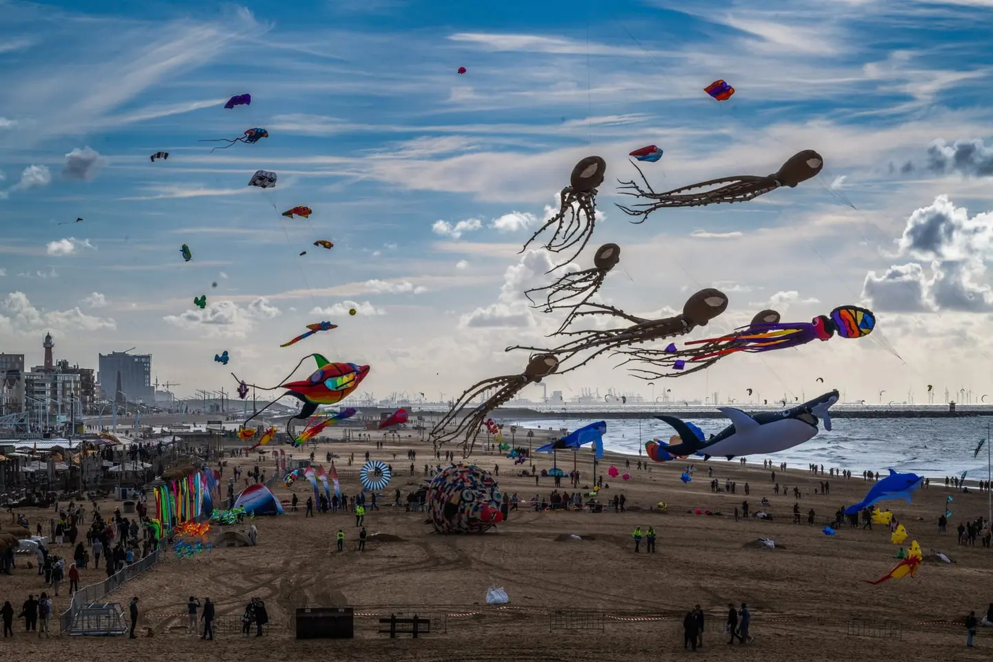A panoramic beachscape teems with crowds and colorful kites of all sizes — octopuses, whales, and abstract shapes — drifting beneath a dramatic streaked sky above an industrial harbor skyline.