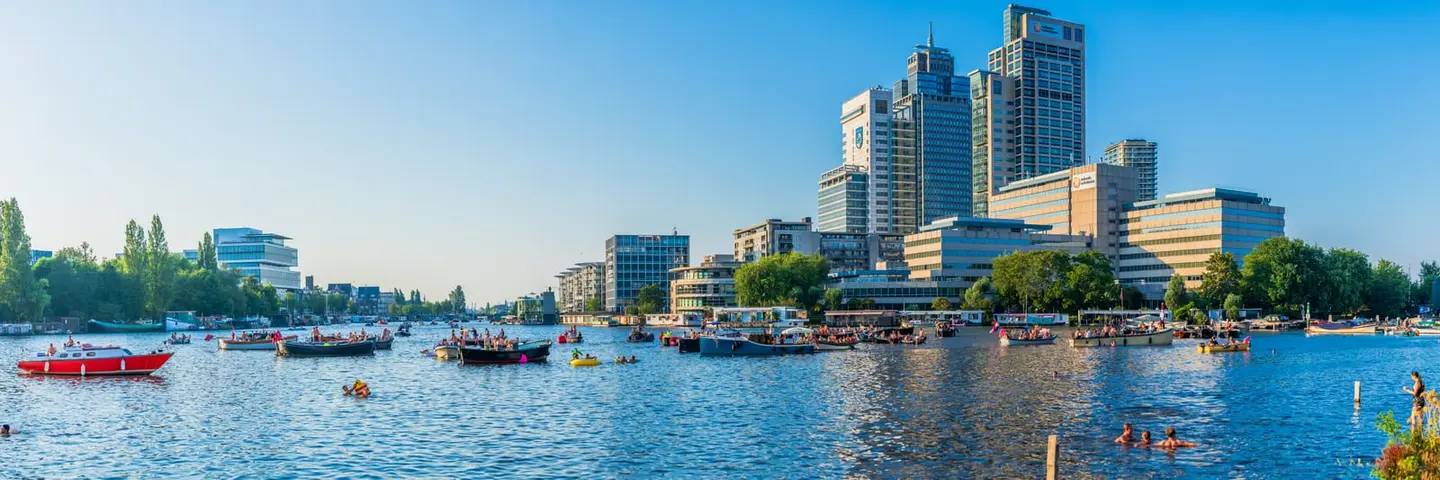 A summer evening on the Amstel in Amsterdam Oost — dozens of small boats, sloops and inflatables drift along the broad river while swimmers cool off in the foreground.
