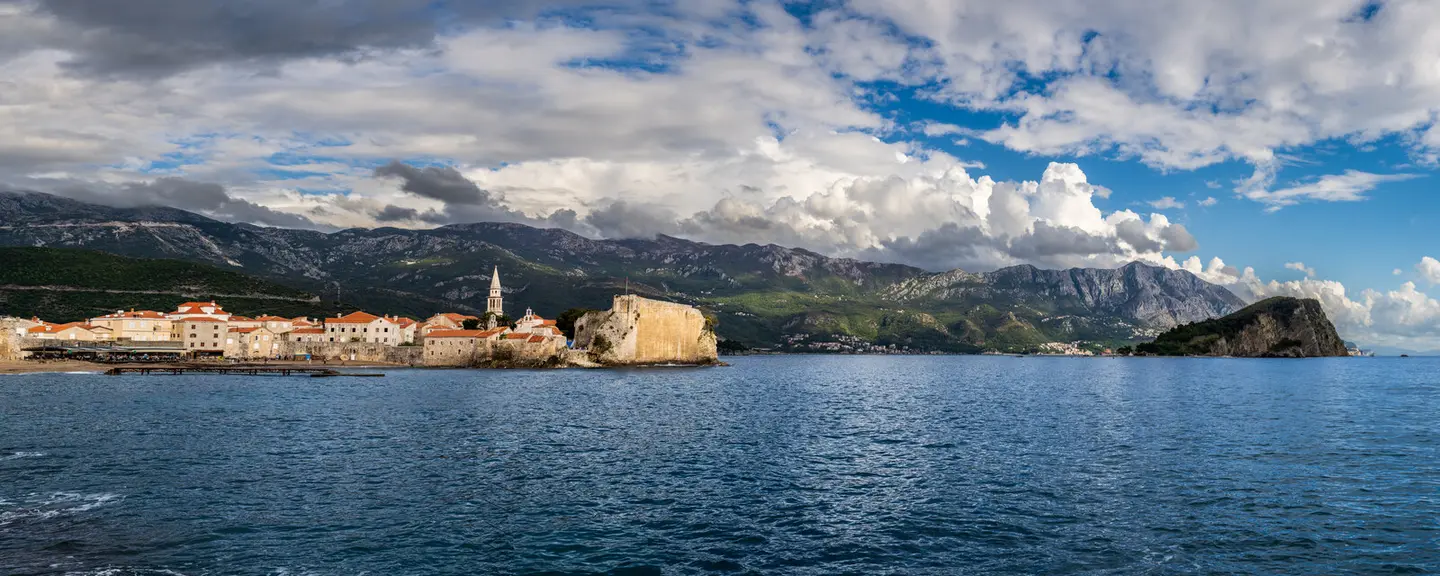 Budva's walled old town juts into the Adriatic on its rocky peninsula, terracotta roofs and the stone campanile catching afternoon light beneath a sweeping bank of cumulus cloud.