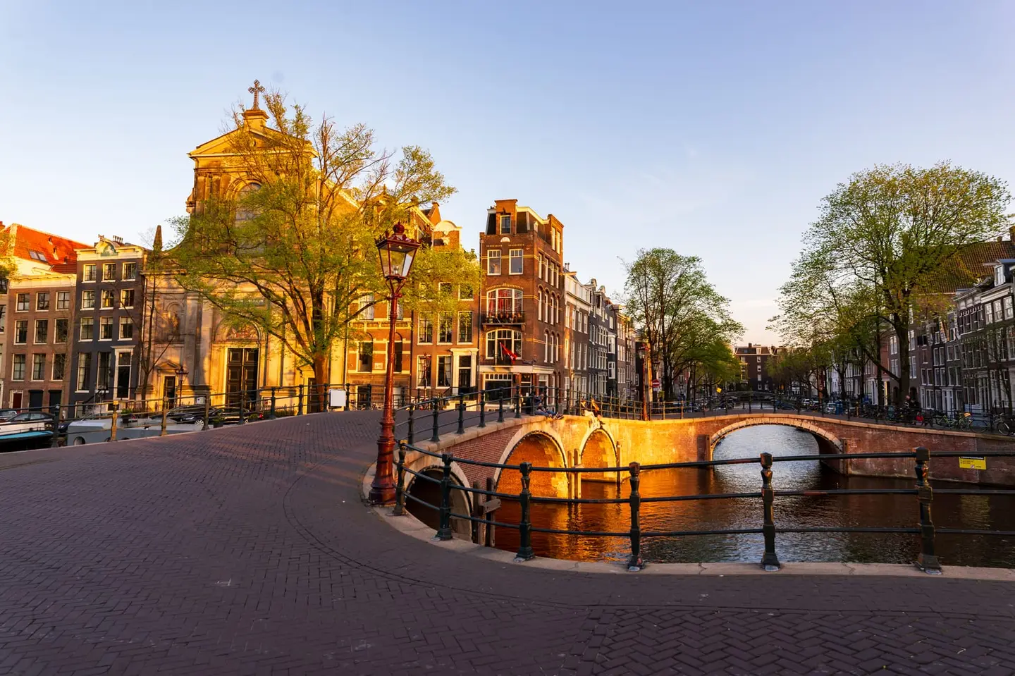 Warm evening sun floods a curving brick quay in Amsterdam, leading the eye to a low arched canal bridge and the twin-towered facade of the Posthoornkerk beyond.