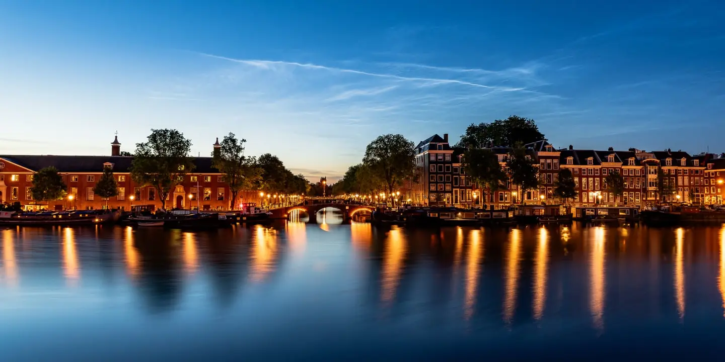 Blue hour settles over the river Amstel in Amsterdam, where a long classical facade and a row of gabled canal houses line the far bank beneath a sweep of wispy high cloud.